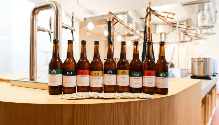 Huit bouteilles de bière artisanale alignées, chacune avec une étiquette colorée indiquant son nom et le degré alcool. Un tireuse à bière est visible à gauche.