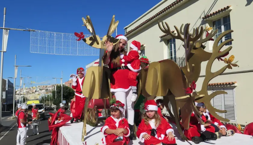Niños y adultos disfrazados de Papá Noel y elfos participan en una cabalgata de Papá Noel, con un gran reno dorado y decoraciones navideñas.