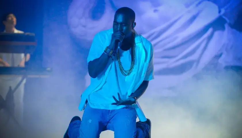 Kanye West na koncercie, klęczy z mikrofonem, otoczony niebieskim światłem i mgłą.