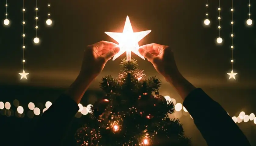 Hände setzen einen leuchtenden Stern auf die Spitze eines Weihnachtsbaums, der die weihnachtliche christliche Bedeutung des Lichts und der Hoffnung symbolisiert.