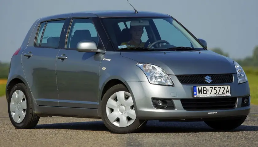 Szary Suzuki Swift 2007 z kierowcą za kierownicą, zaparkowany na drodze.