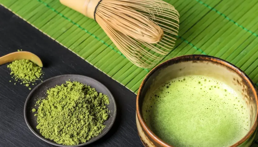 Matcha-Pulver auf einem Teller, daneben eine Schale mit schaumiger Matcha-Creme und ein Bambusbesen.