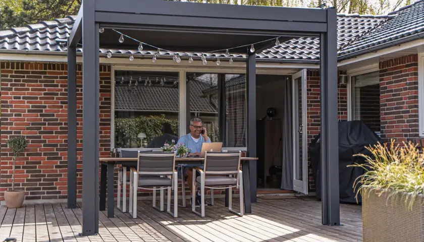 Man arbetar på laptop vid ett bord under en pergola, perfekt för att bygga uteplats. Ljusslinga ovanför.