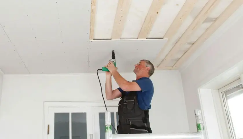 Man på byggställning skruvar gipsskivor i taket. Taket är under renovering med synliga träreglar.