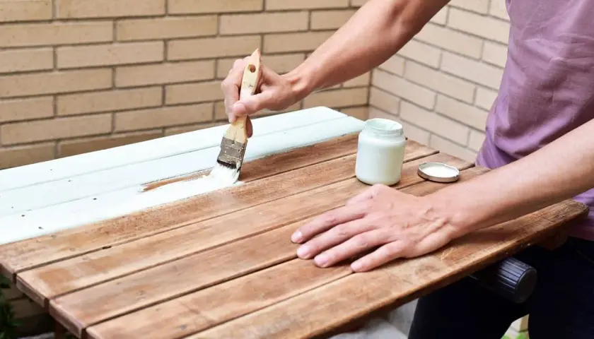Persona pintando una mesa de madera exterior con brocha y pintura blanca.