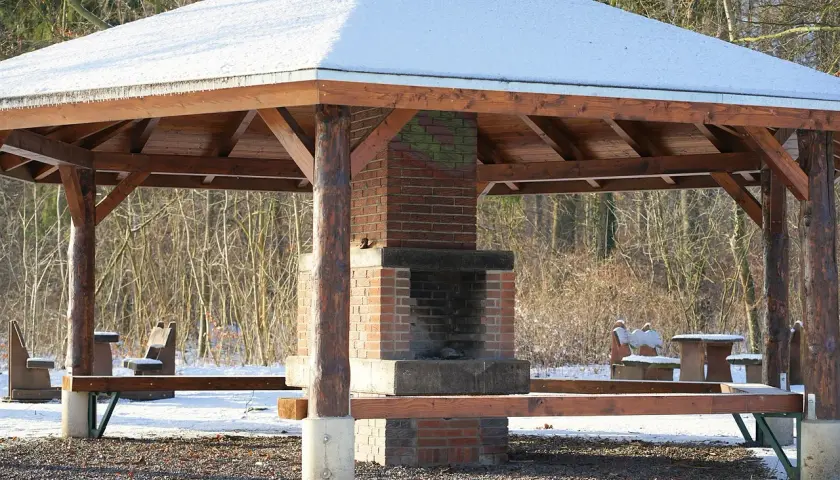 En träpaviljong med snö på taket och en murad grill i mitten. Bänkar och bord står redo för en mysig fika utomhus.