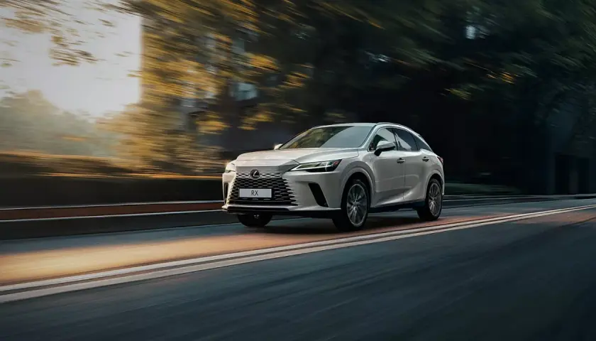 Biały Lexus RX 350h pędzi po drodze, otoczony rozmazanymi drzewami i światłem zachodzącego słońca.