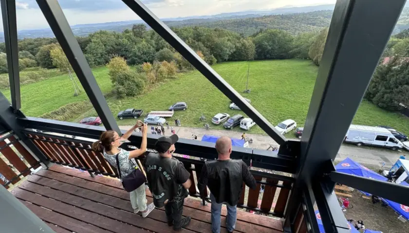 Widok z wieży widokowej na malowniczy krajobraz z lasami i polami. Na dole widać zaparkowane samochody, namioty i ludzi, jakby szykowali się na festyn lub zlot. Może to być nawet zlot motocyklowy, bo jeden z panów ma na sobie kamizelkę z napisem "GÓRY ...