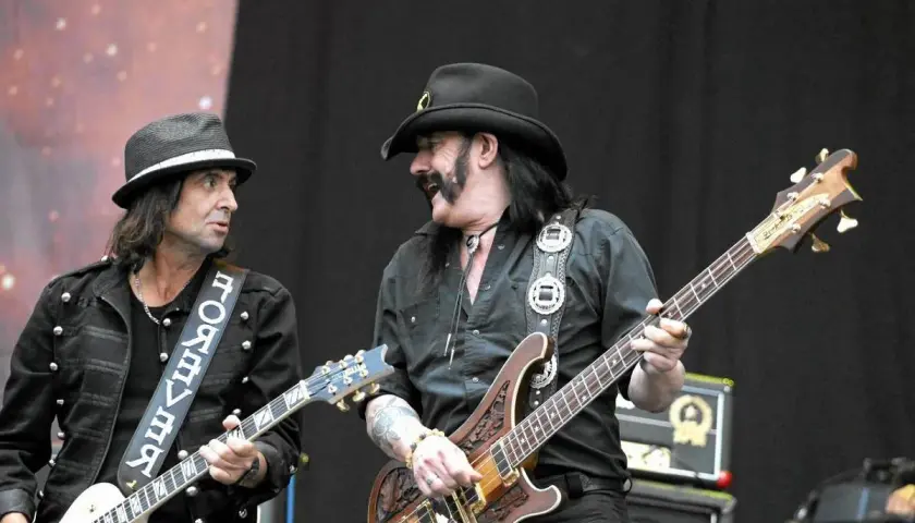 Lemmy Kilmister z zespołem na scenie. Dwóch muzyków gra na gitarach, jeden w kapeluszu, drugi w kowbojskim.