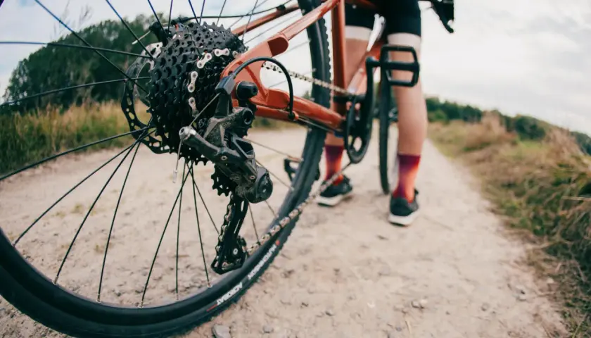 Widok z bliska tylnej przerzutki rowerowej, która wygląda na Shimano Acera lub Alivio, na tle szutrowej drogi i rowerzysty.