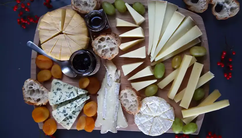 Un ricco tagliere con vari formaggi da aperitivo, pane croccante, uva, albicocche secche e marmellata.