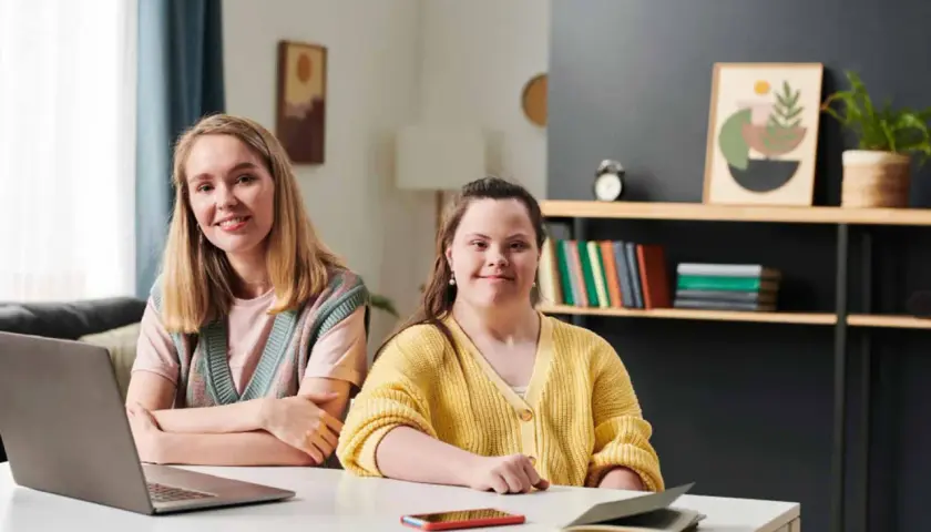 Dwie kobiety siedzą przy biurku, jedna z zespołem Downa, obie uśmiechnięte.