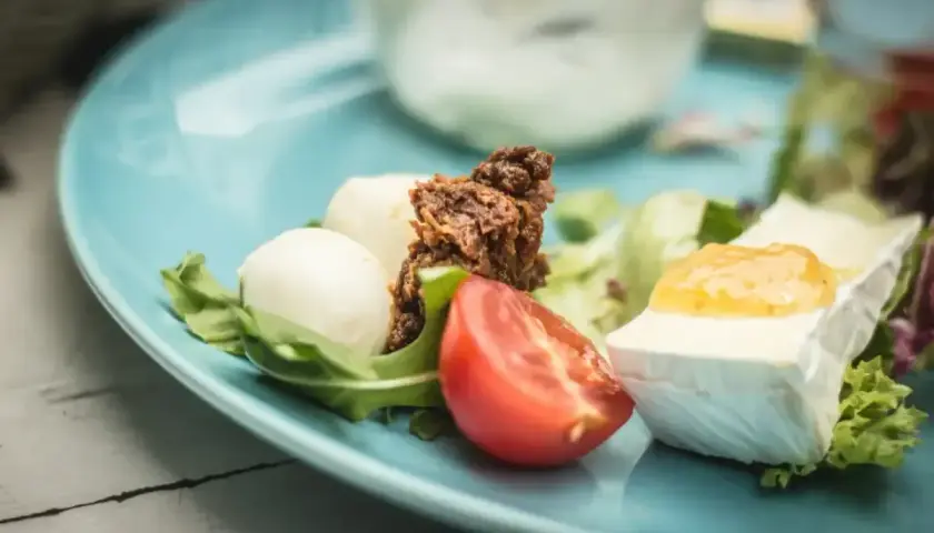 Keto śniadanie: ser brie z dżemem, kulki mozzarelli, pomidor i rukola na niebieskim talerzu.