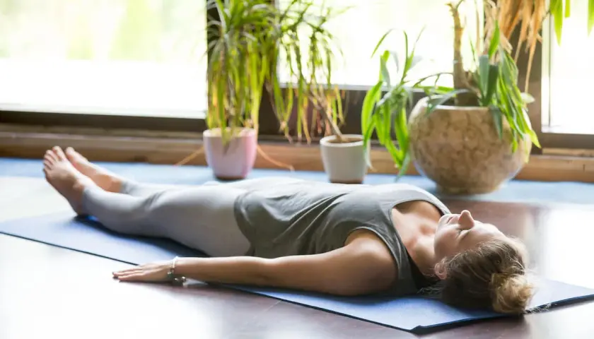 Kobieta w pozycji savasana, zrelaksowana na macie do jogi, otoczona zielenią roślin.