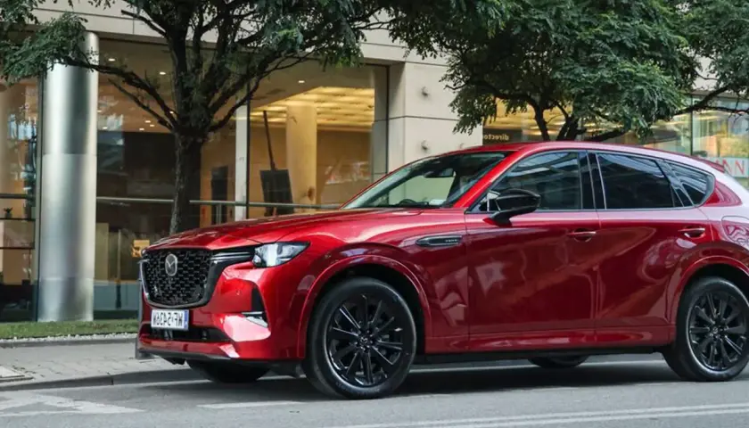 Lśniąca, czerwona Mazda CX-60 3.3 diesel na tle nowoczesnego budynku i drzew.