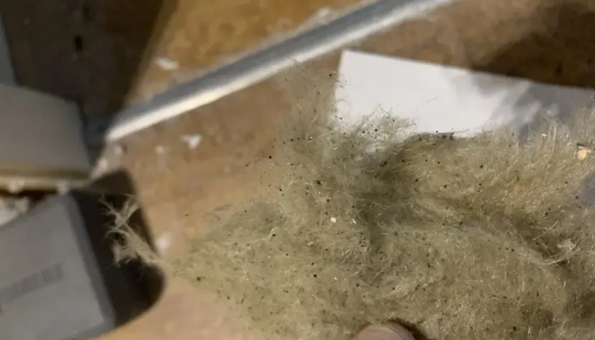 En hand håller en tuss av stenull eller glasull, som är dammig och innehåller små svarta prickar.