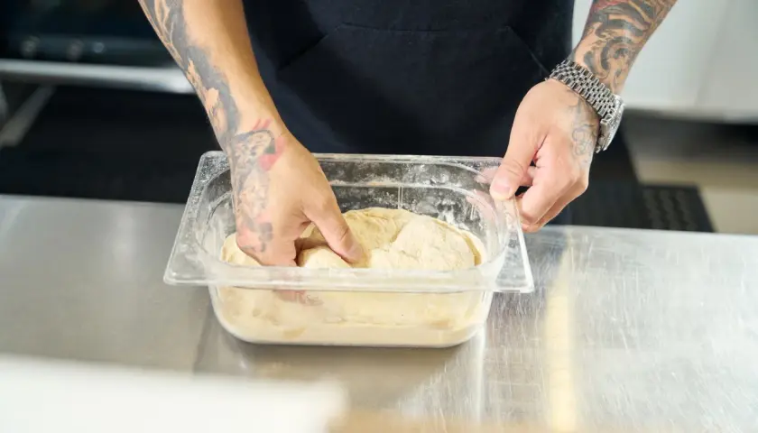 Ręce z tatuażami wyjmują ciasto na pizzę z pojemnika. Dowiedz się, jak rozmrozić ciasto na pizzę, aby uzyskać idealną bazę.