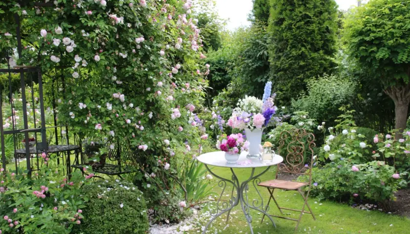 Ein romantischer Garten mit blühenden Rosen, ein Tisch mit Blumenarrangements und ein schmiedeeiserner Stuhl laden zum Verweilen ein. Ein idyllischer Ort, um einen **beet anlegen**-Traum zu verwirklichen.