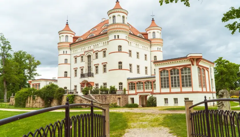 Zamek w Łomnicy, biała budowla z czerwonymi dachami i wieżyczkami, otoczona zielenią.