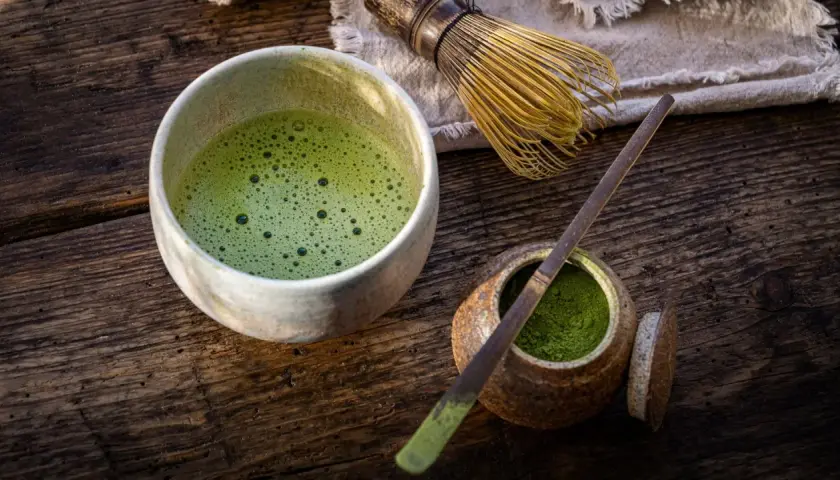 Un bol de matcha mousseux, une cuillère en bambou et de la poudre verte dans un pot sur une table en bois.
