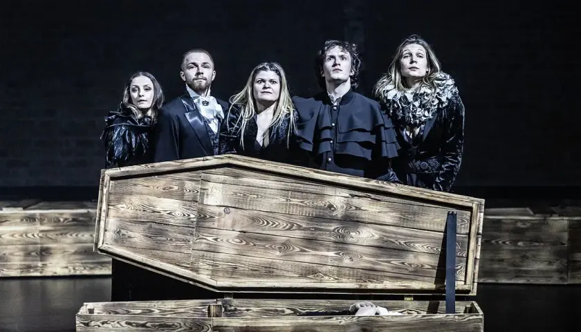 Aktorzy Teatru Pantomimy Wrocław w strojach z epoki stoją nad otwartą trumną, z której wystaje ręka.