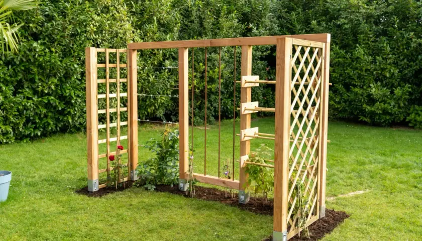 Holzkonstruktion im Garten mit Rankhilfen, die an Baustahlmatten erinnern. Kletterrosen und junge Pflanzen wachsen daran empor.