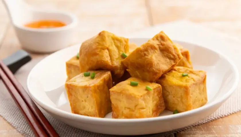 Tofu crujiente dorado, cortado en cubos y adornado con cebollín, servido en un plato blanco con salsa y palillos.
