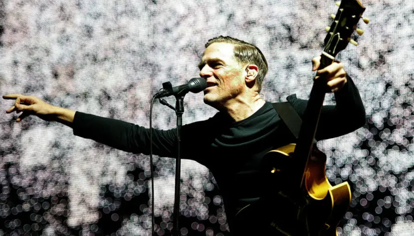 Bryan Adams na koncercie, śpiewa do mikrofonu i gra na gitarze.