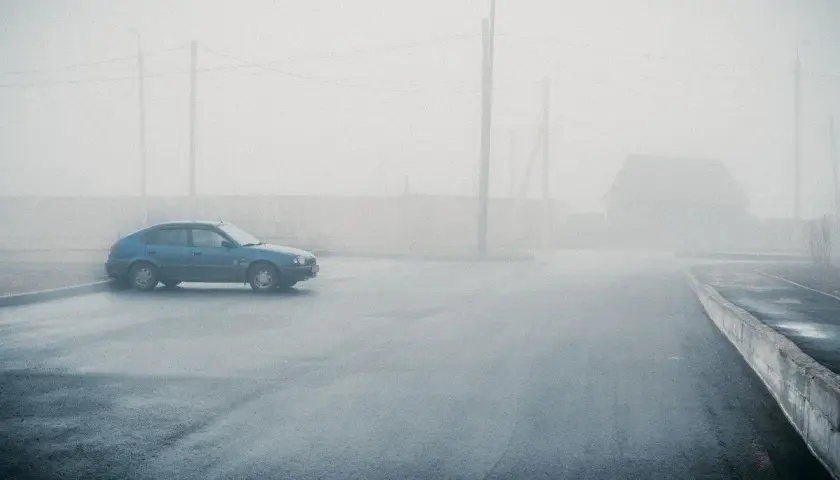 Un coche azul solitario se detiene en medio de una **niebla espesa** que oculta un camino y una casa.