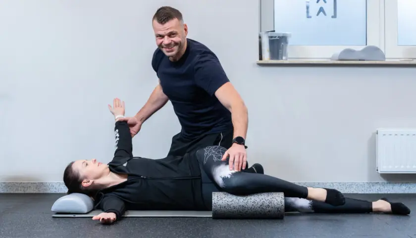 Fizjoterapeuta pomaga pacjentce w ćwiczeniach na kręgosłup. Kobieta leży na macie, z nogami ułożonymi na wałku, a mężczyzna koryguje jej pozycję ramienia.