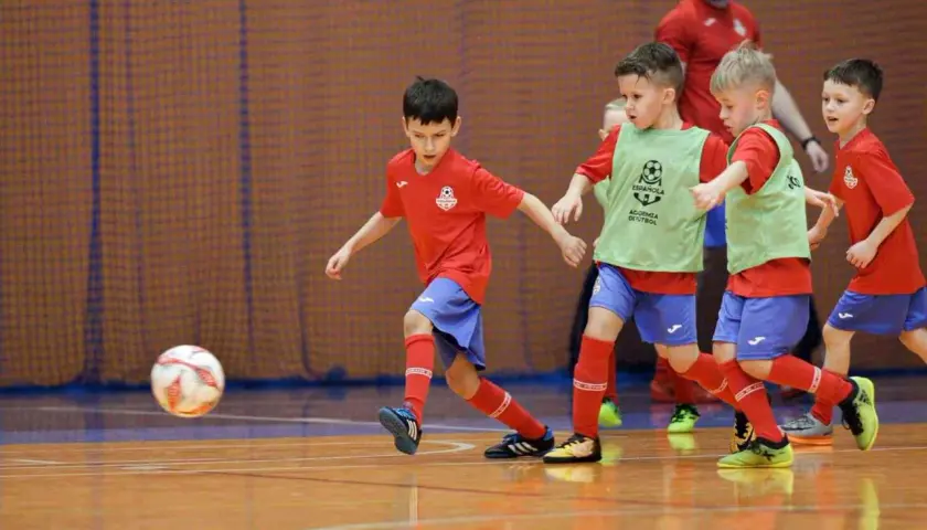 Zacięte współzawodnictwo sportowe dzieci i młodzieży na hali. Mali piłkarze w czerwonych koszulkach i zielonych kamizelkach walczą o piłkę.