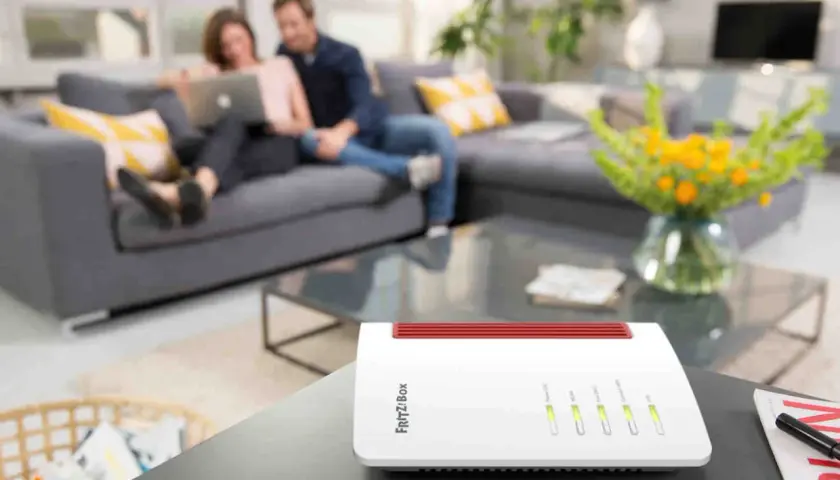 Biały router FRITZ!Box na stoliku, z parą relaksującą się na kanapie w tle. To urządzenie zapewnia domowe Wi-Fi.