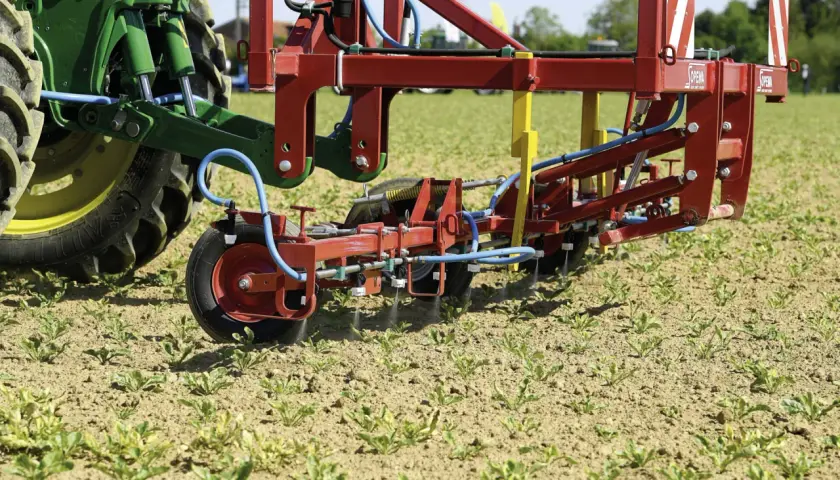 Un tracteur vert et un pulvérisateur rouge travaillent dans un champ de jeunes pousses. La machine applique un traitement ciblé, peut-être pour une liste de graminées à éradiquer.