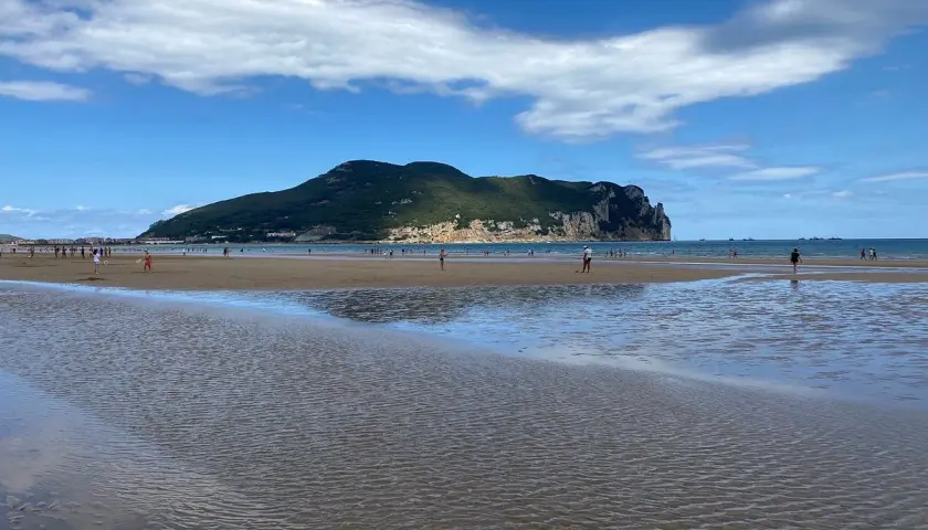 Amplia **playa Laredo** con gente paseando y jugando, bajo un cielo azul con nubes blancas y una gran montaña verde al fondo.
