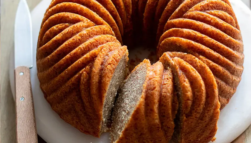 Una torta cachi e mele dorata, con un bordo scanalato, è affettata su un tagliere bianco. Un coltello con manico in legno è appoggiato accanto.