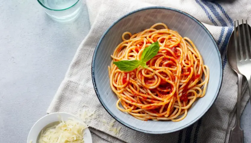 Spaghetti z sosem pomidorowym i bazylią w misce, obok starty ser i sztućce.