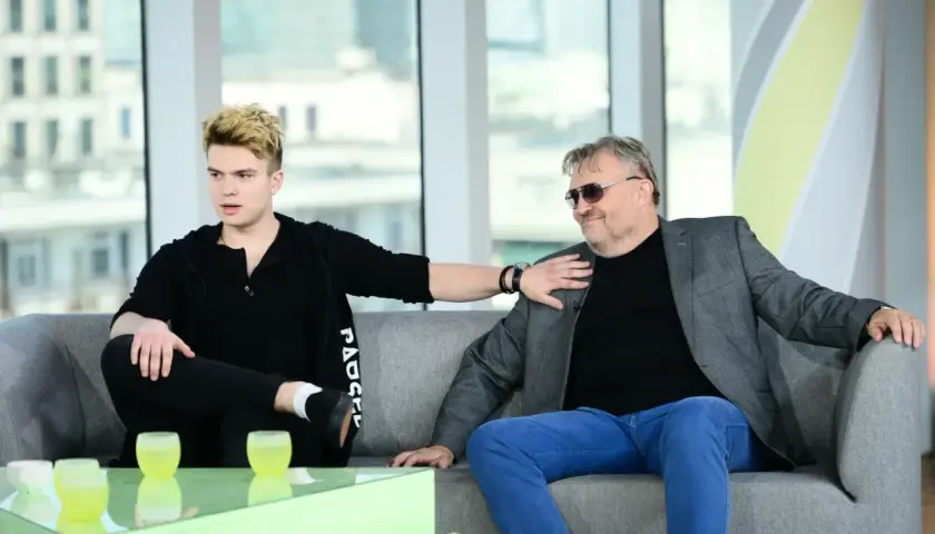 Kris Cugowski i jego syn rozmawiają na kanapie. Młodszy z nich, z blond włosami, gestykuluje, starszy, w okularach, z uśmiechem słucha.