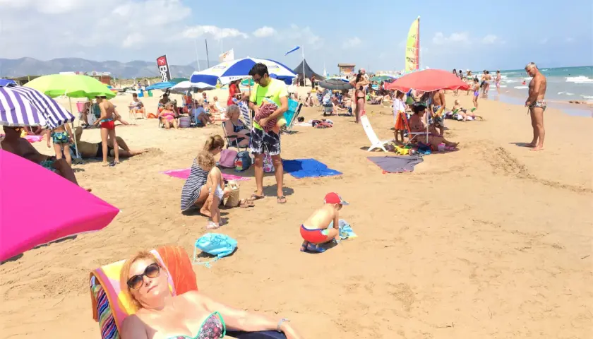 Día soleado en la playa de l'Ahuir, con gente disfrutando bajo sombrillas y el mar al fondo.