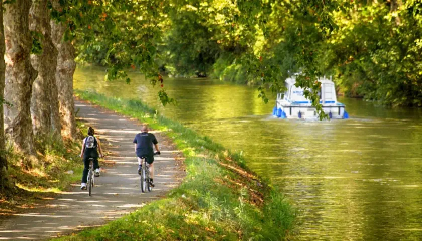 Pareja en bicicleta disfruta de un **canal du midi recorrido** bajo árboles frondosos, mientras una barcaza navega por el agua.