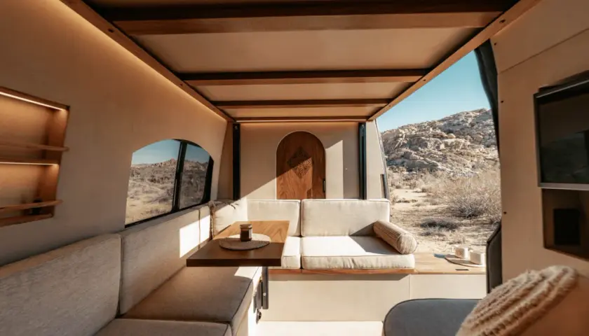 Intérieur de camping-car aménagé avec banquettes, table et porte en bois, vue sur un paysage désertique.