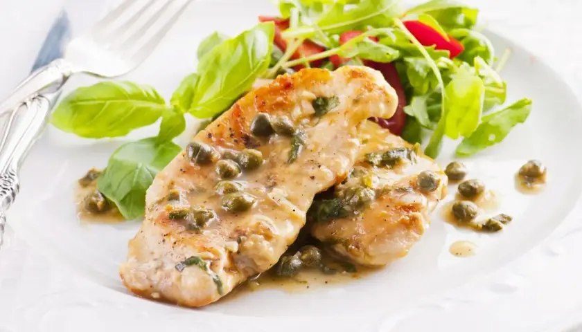 Deliziose scaloppine di pollo al limone e capperi, servite con un'insalata fresca e foglie di basilico.