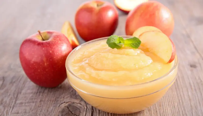 Un tazón de deliciosa salsa de manzana casera, decorado con menta y rodajas de manzana, rodeado de manzanas rojas frescas sobre una mesa de madera.