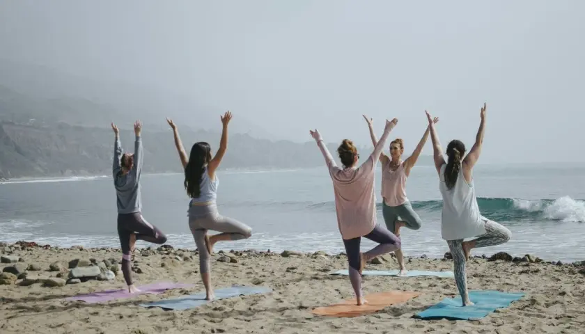 Fem kvinnor utför pilates yoga på stranden med havet i bakgrunden.