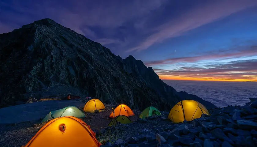 Acampada en la montaña al amanecer. Tiendas iluminadas sobre rocas, con un mar de nubes y cielo anaranjado. ¡Espero que no haya multa por acampar aquí!