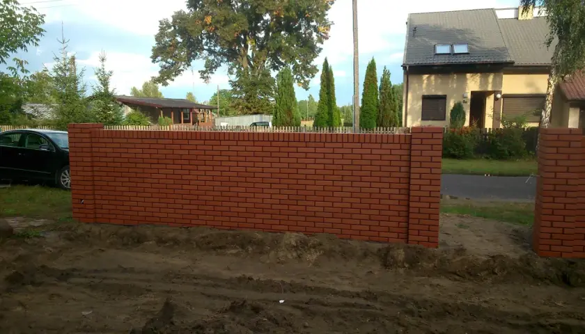 Nowy, ceglany szamot otacza posesję. W tle dom i drzewa.