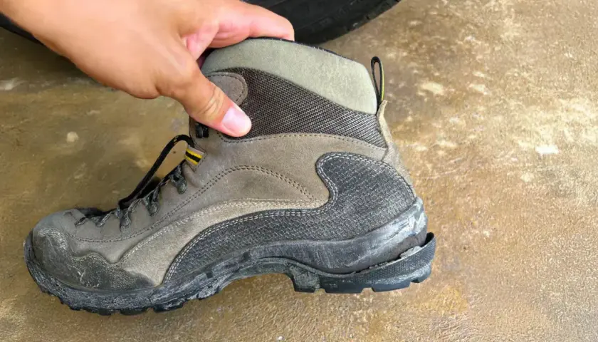 A hand holds a worn hiking boot, showing why shoe soles crack: exposure to elements and heavy use can cause the rubber to degrade and split.