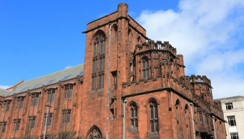 Imponująca, ceglana architektura w stylu gotyckim w Manchesterze, z charakterystycznymi wieżyczkami i łukowymi oknami.
