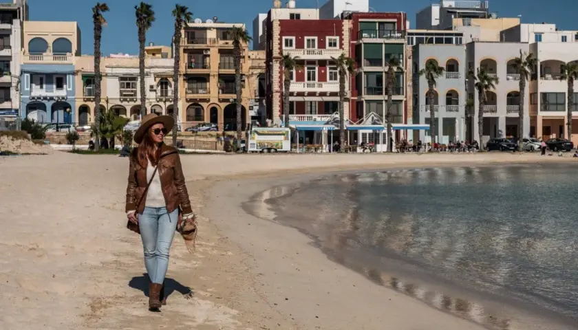 Kobieta w kapeluszu spaceruje po piaszczystej plaży z kolorowymi budynkami w tle.
