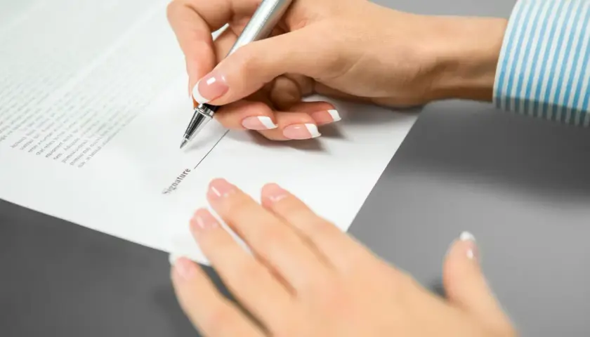 Mano femenina con uñas cuidadas escribe en un documento. El texto "Signature" es visible, pero el **contrato sin firmar** carece de **validez** hasta que se complete.