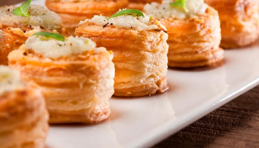 Petites bouchées à la reine dorées, garnies d'une préparation crémeuse et d'une feuille de basilic, présentées sur un plat blanc.